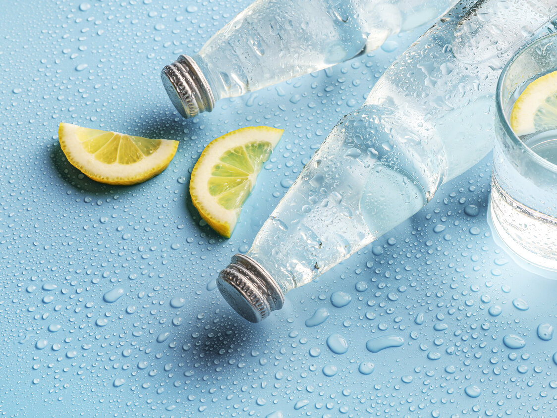 Zwei Flaschen Wasser und ein Glas mit Wasser auf blauem Untergrund. Daneben Zitronen.