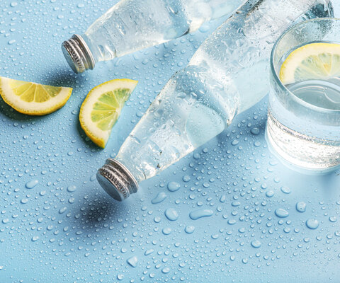 Zwei Flaschen Wasser und ein Glas mit Wasser auf blauem Untergrund. Daneben Zitronen.