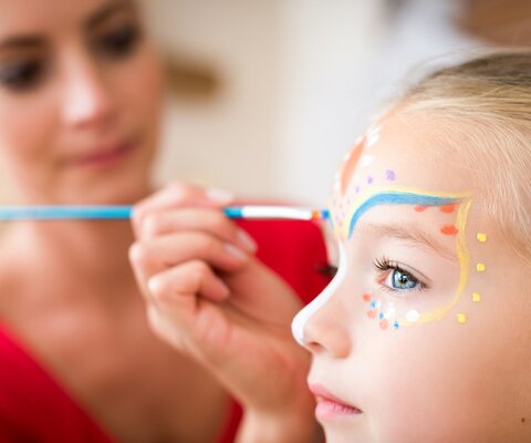 Eine Frau malt mit einem Pinsel das Gesicht eines Kindes an.