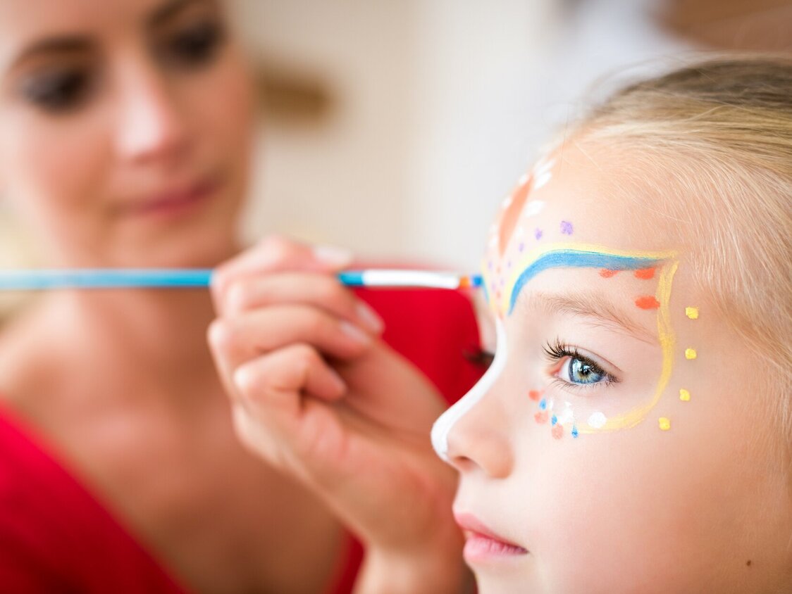 Eine Frau malt mit einem Pinsel das Gesicht eines Kindes an.