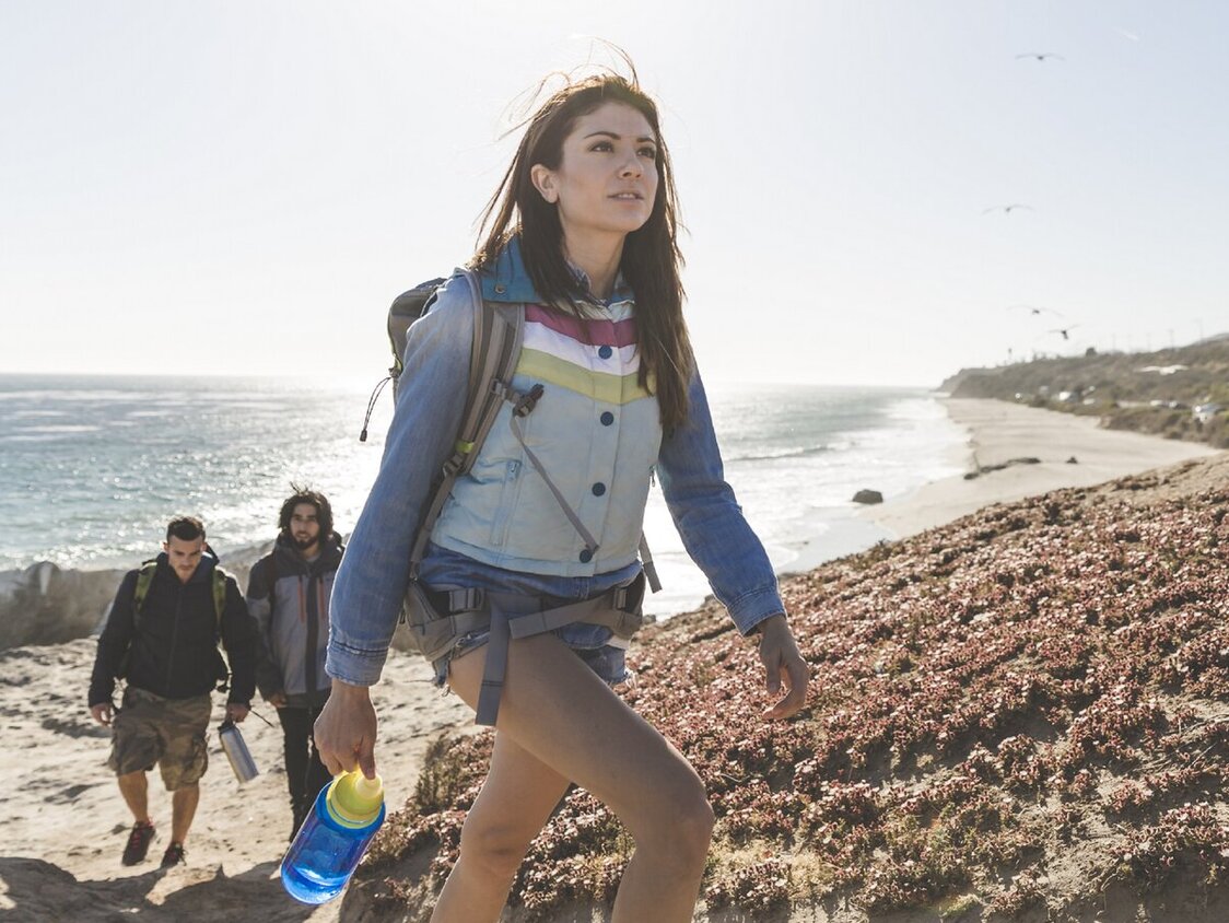  Eine Frau und zwei Männer, die einen Berg an einem Strand hinauf laufen. Im Hintergrund das Meer, blauer Himmel und Sonne.