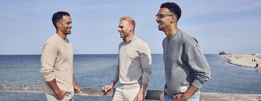 Drei Männer stehen auf einem Steg am Meer und unterhalten sich. Sie lehnen am Geländer, lachen und sind gut gelaunt. Sie haben ihre Hände in der Hosentasche. Die Sonne scheint und der Himmel ist blau.