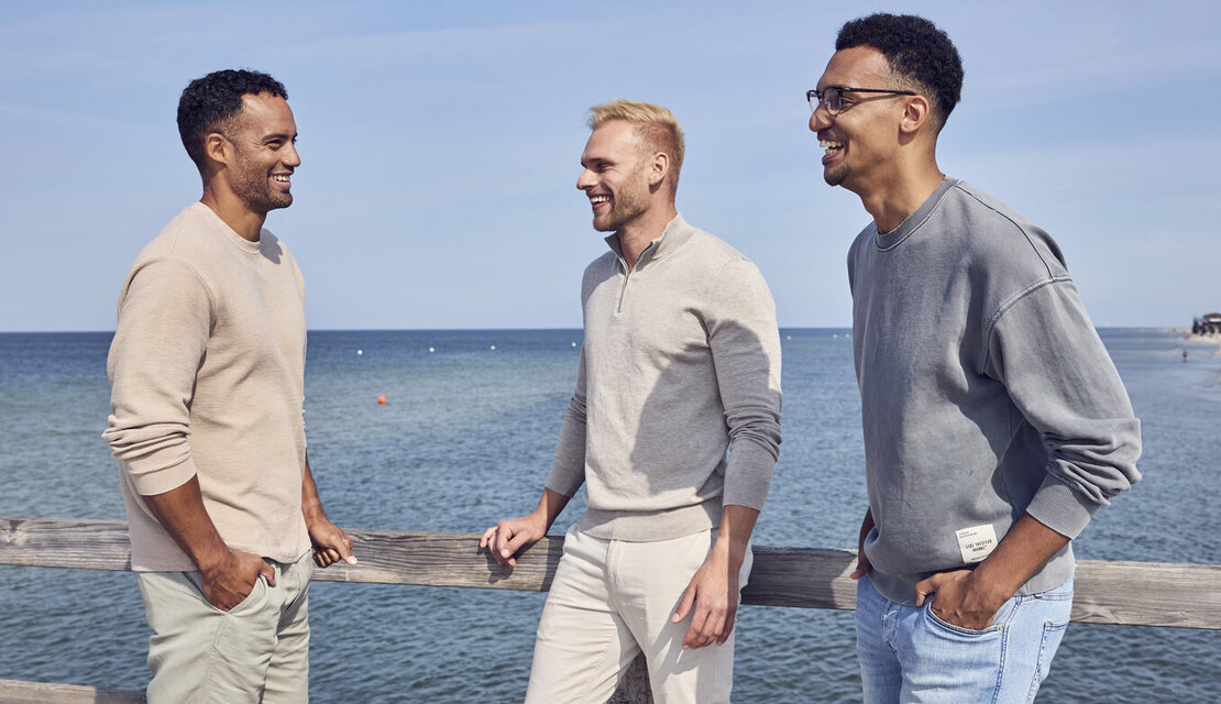 Drei Männer stehen auf einem Steg am Meer und unterhalten sich. Sie lehnen am Geländer, lachen und sind gut gelaunt. Sie haben ihre Hände in der Hosentasche. Die Sonne scheint und der Himmel ist blau.