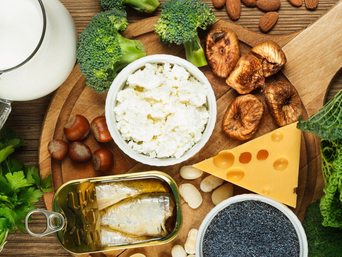 Auf einem Holzbrett liegen einige kalziumreiche Lebensmittel wie z.B. eine Kanne Milch, eine Dose Sardinen, ein Stück Käse, einige Mandeln und gerocknete Feigen, sowie Brokkoli und Wirsing.