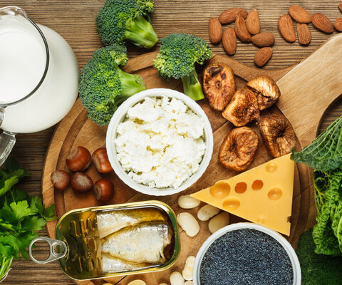 Auf einem Holzbrett liegen einige kalziumreiche Lebensmittel wie z.B. eine Kanne Milch, eine Dose Sardinen, ein Stück Käse, einige Mandeln und gerocknete Feigen, sowie Brokkoli und Wirsing.