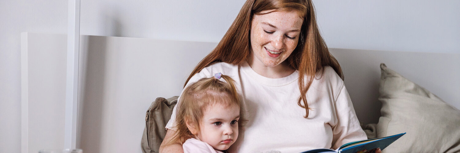 Eine Mutter sitzt mit ihrem Kind im Bett und liest ihm aus einem Buch vor. Auf dem Nachttisch steht Medizin.