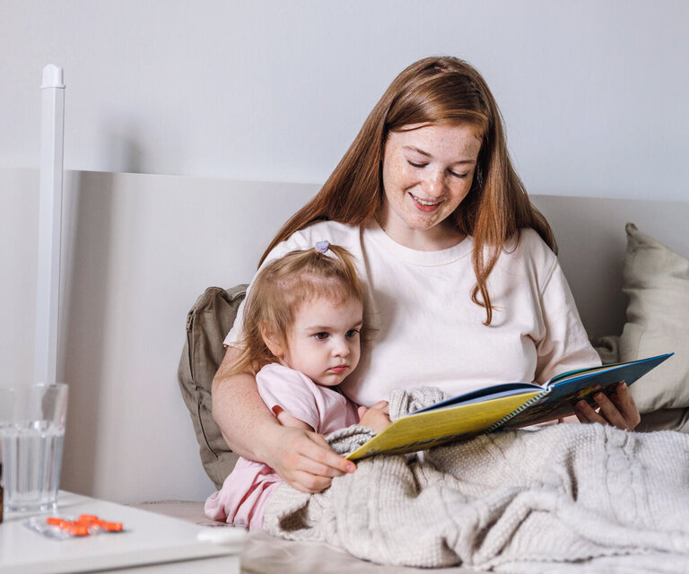 Eine Mutter sitzt mit ihrem Kind im Bett und liest ihm aus einem Buch vor. Auf dem Nachttisch steht Medizin.