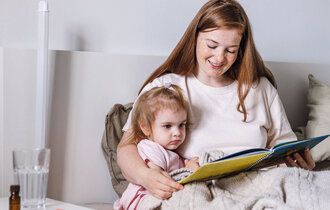 Eine Mutter sitzt mit ihrem Kind im Bett und liest ihm aus einem Buch vor. Auf dem Nachttisch steht Medizin.