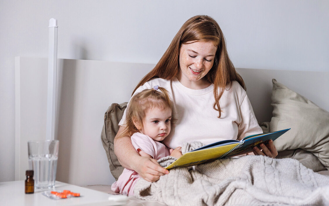 Eine Mutter sitzt mit ihrem Kind im Bett und liest ihm aus einem Buch vor. Auf dem Nachttisch steht Medizin.
