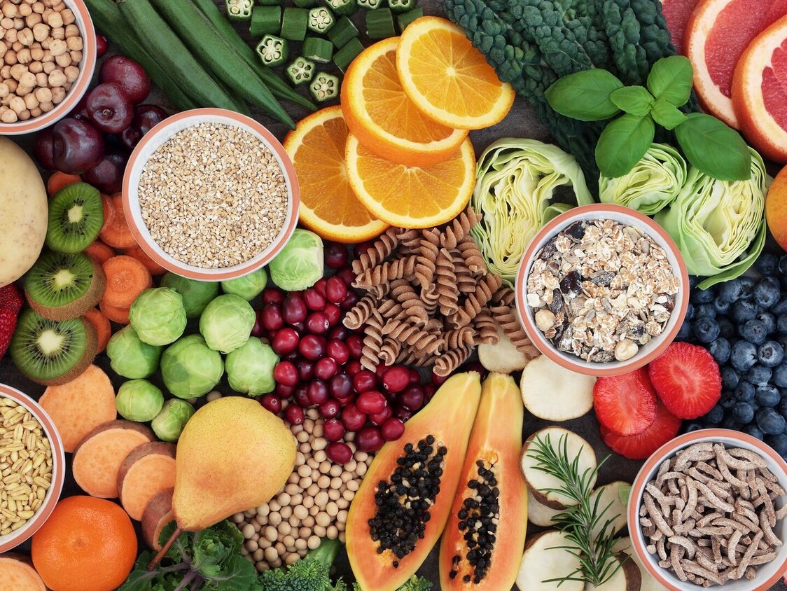  Aufnahme von oben. Auf einem Tisch liegen gesunde Lebensmittel: Obst, Vollkornnudeln, Nüsse, Blaubeeren, Apfel, Orangen, Kiwi, Erdbeeren, Birne, Melone, Pflaume, Brokkoli, Mandarine, Grapefruit, Rosenkohl.