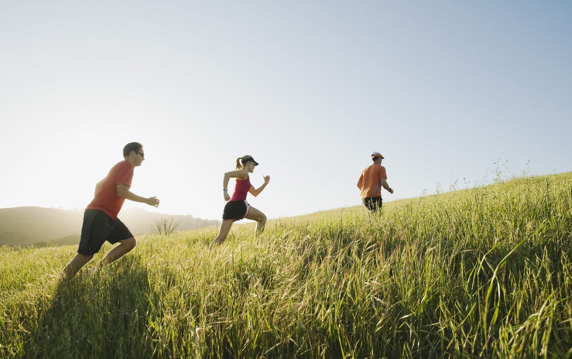 Eine Frau und zwei Männer in Sportkleidung joggen auf einem Pfad durch ein Getreidefeld.