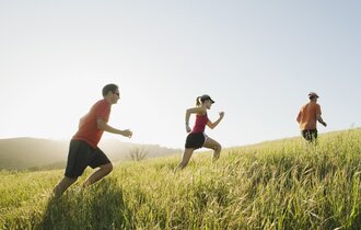 Eine Frau und zwei Männer in Sportkleidung joggen auf einem Pfad durch ein Getreidefeld.