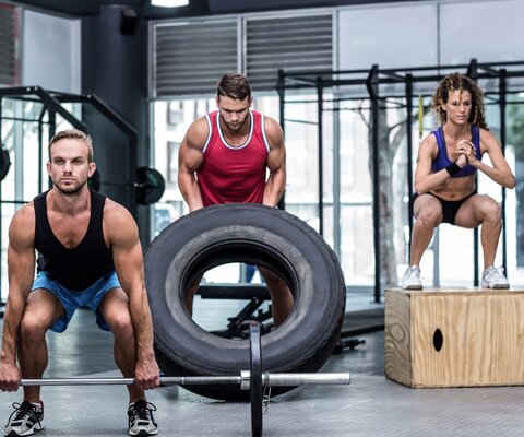 Ein Mann hebt eine Langhantel, ein anderer einen Reifen und eine Frau macht eine Kniebeuge auf einer Box.