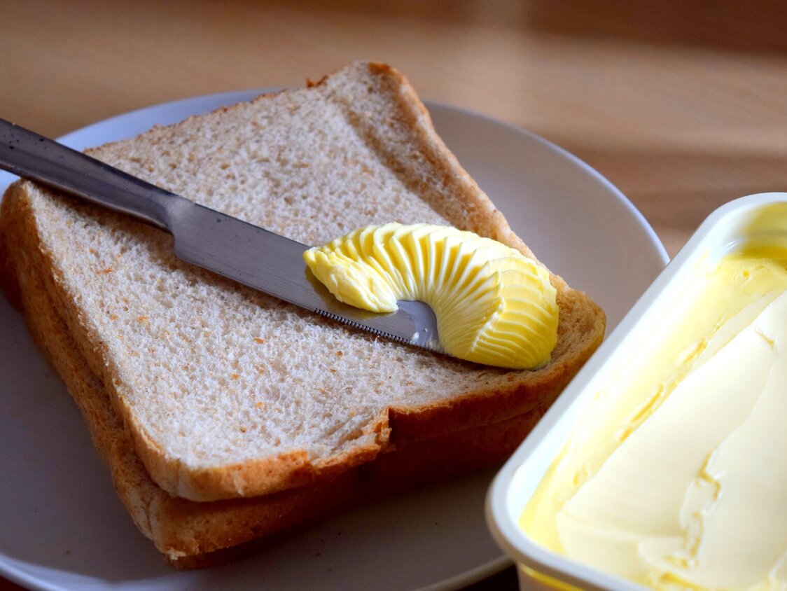 Eine offene Packung Magarine steht neben einem Toastbrot. Auf dem Toast liegt ein Messer mit Butter.