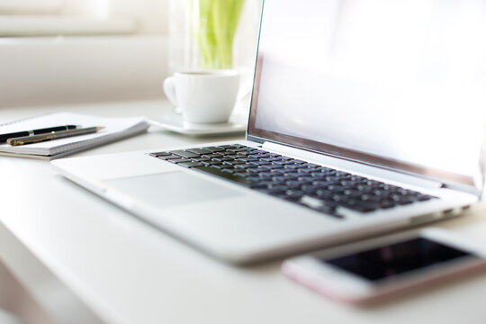 Ein aufgeklappter Laptop steht auf einem Tisch. Daneben liegen ein Block mit Stiften, eine weiße Tasse und ein Smartphone.