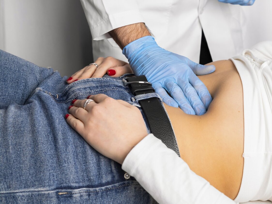 Ein Arzt tastet mit blauen Handschuhen den Bauch einer Frau ab.