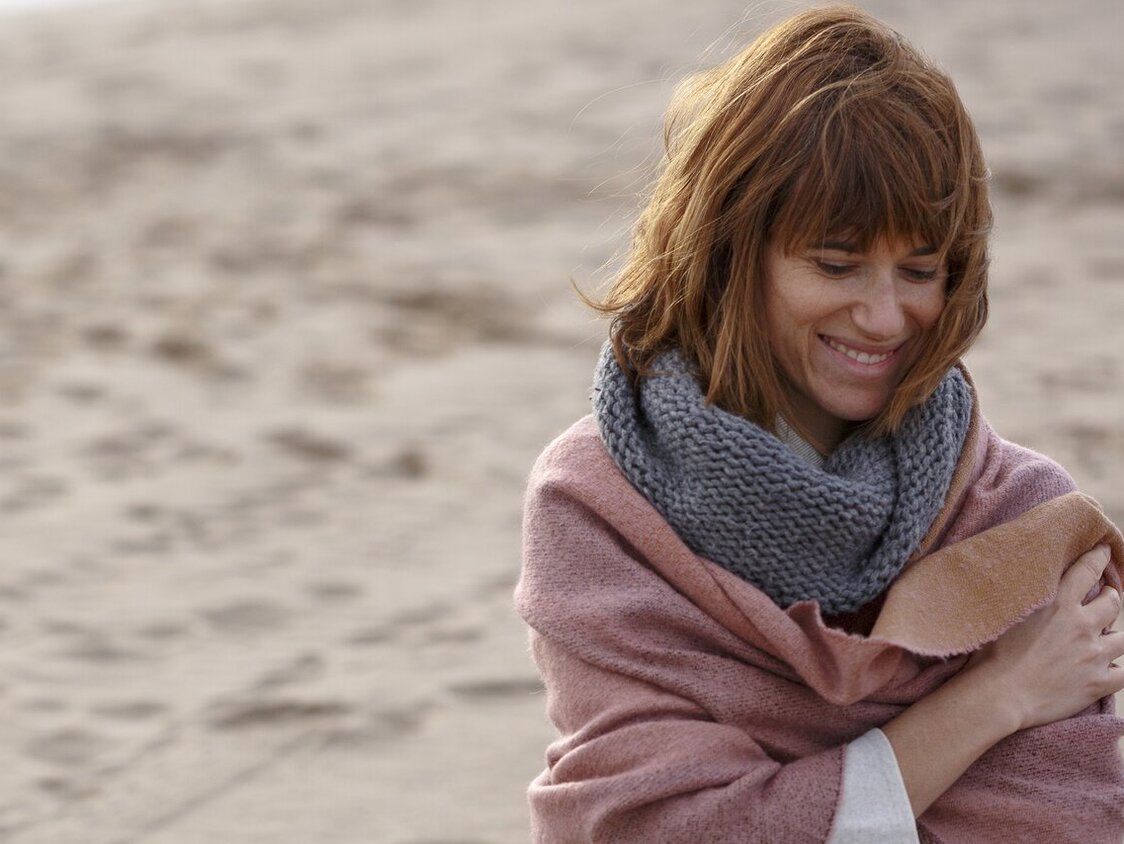 Am STrand steht eine Frau, die sich einen rosa Schal um die Schultern wickelt.