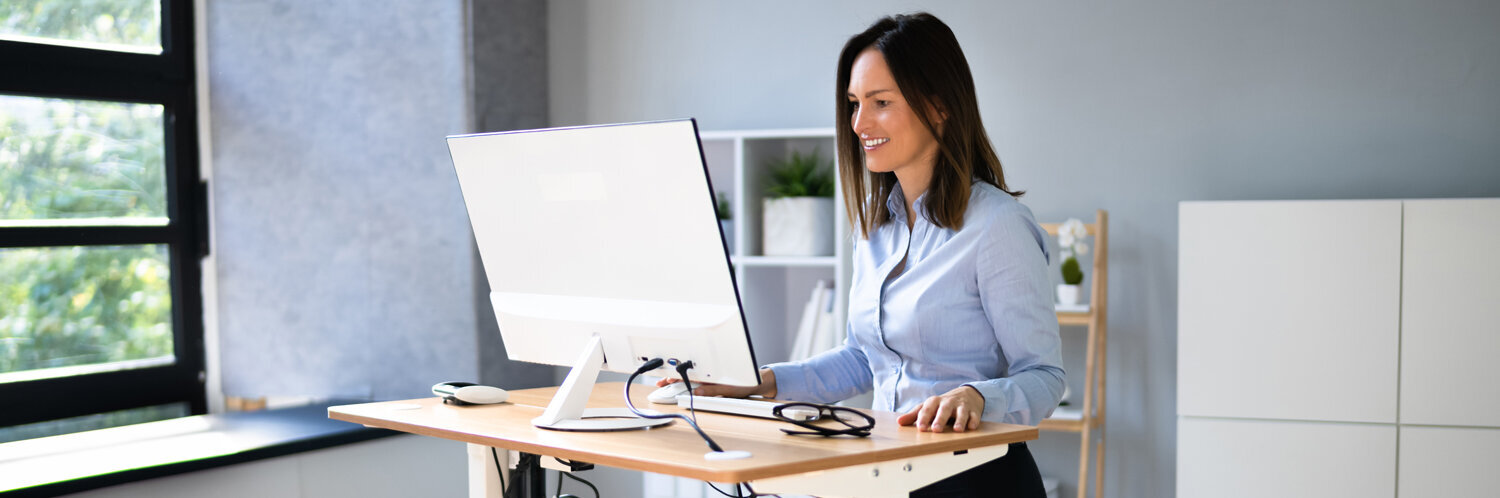Eine Frau steht an einem höhenverstellbaren Schreibtisch und arbeitet an ihrem Computer.
