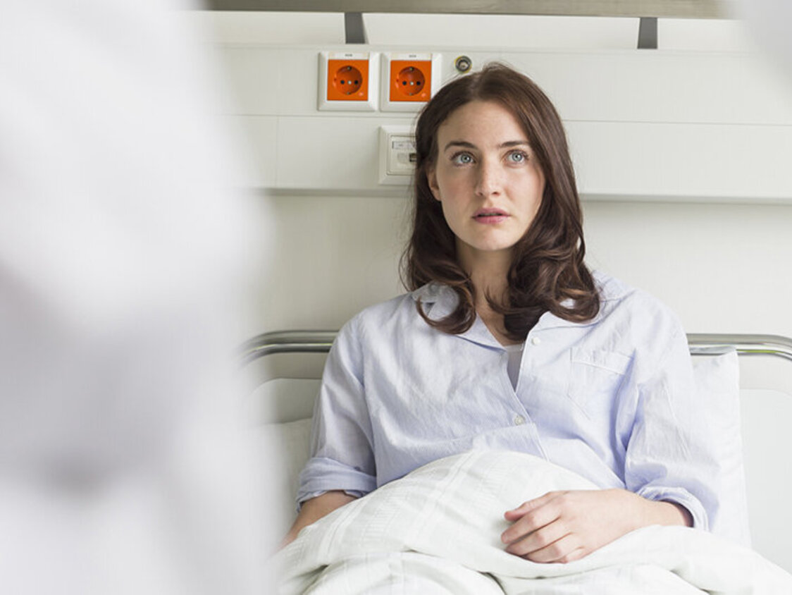 Eine Frau liegt im Krankenhaus in einem Bett und schaut ängstlich zwei Ärzte an.