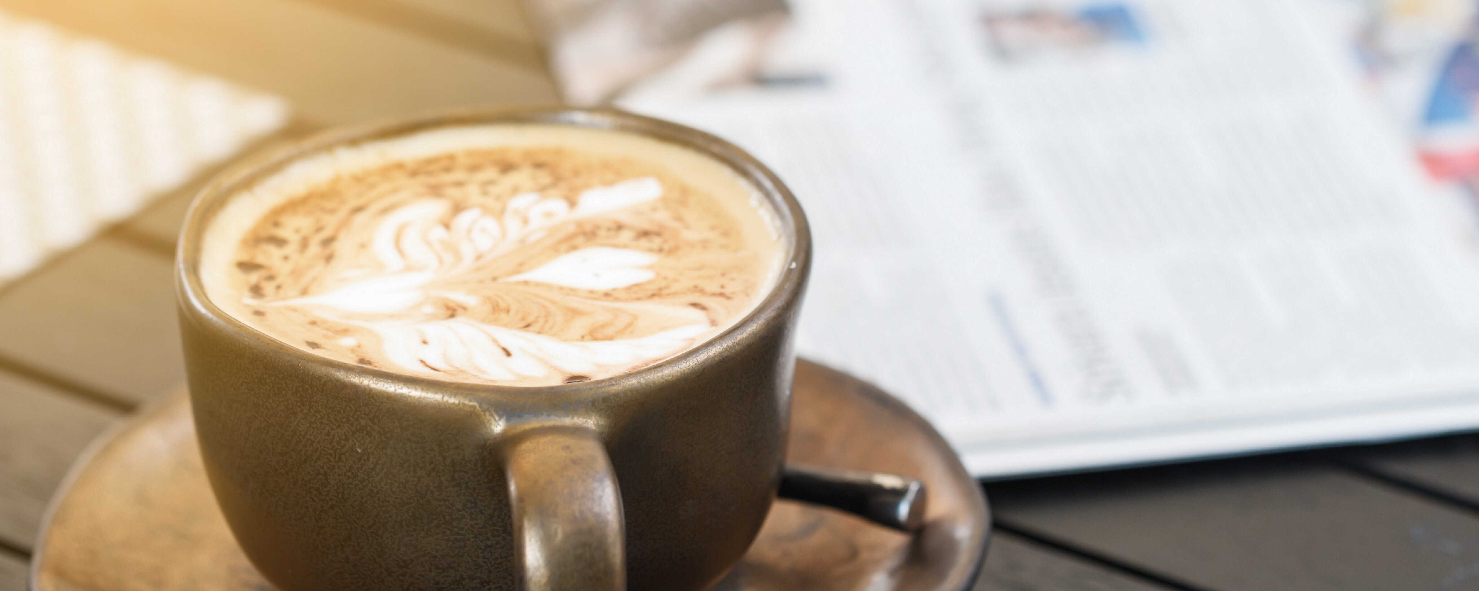 Eine volle Kaffeetasse steht neben einer Zeitung auf einem Tisch.