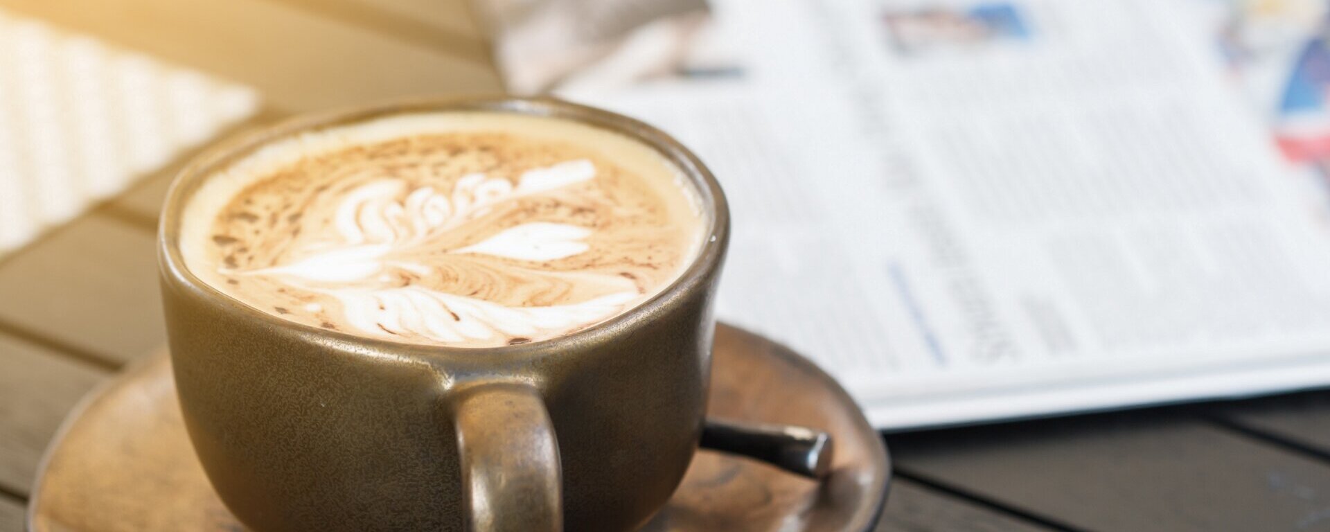 Eine volle Kaffeetasse steht neben einer Zeitung auf einem Tisch.