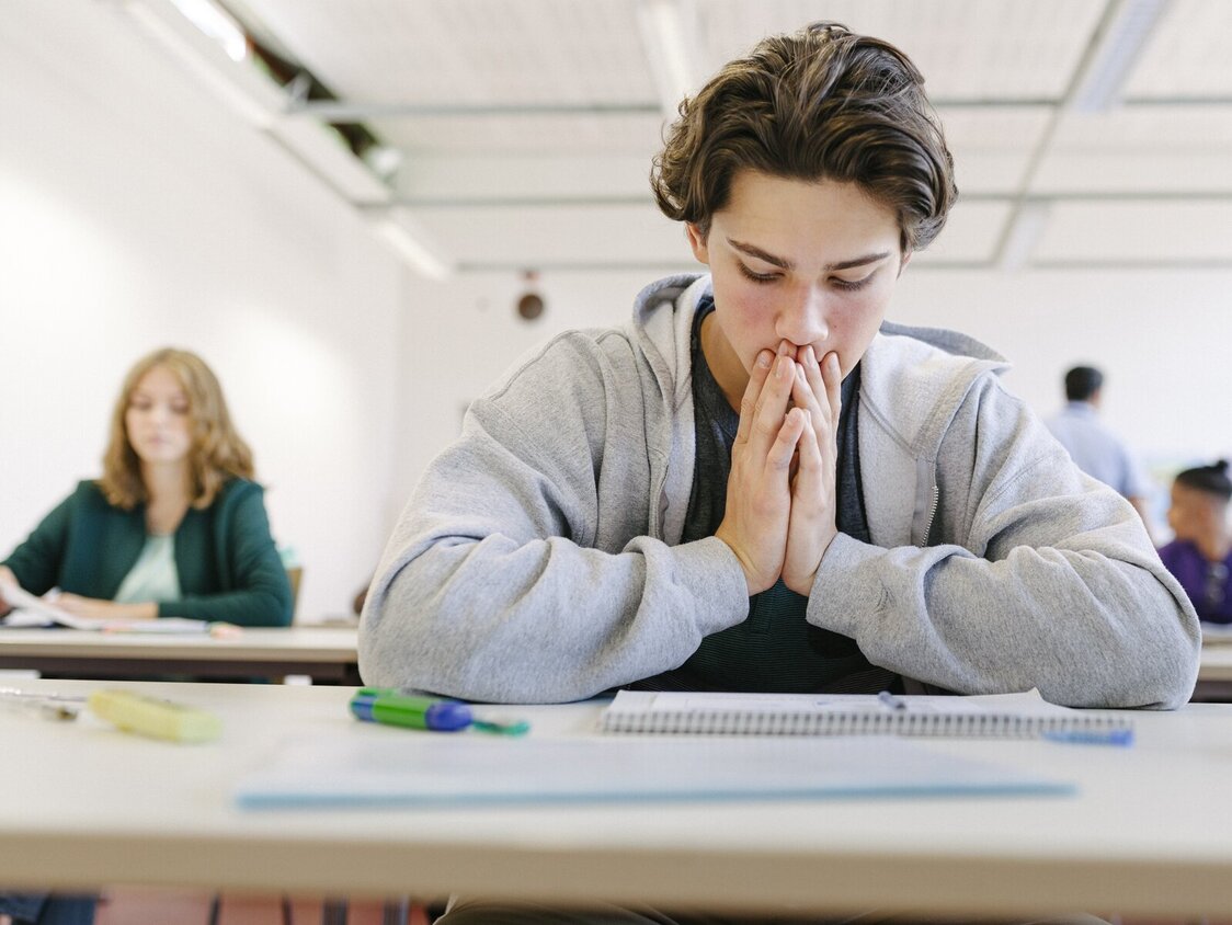 Ein Junge sitzt in der Schule am Tisch und guckt verzweifelt auf seine Unterlagen.