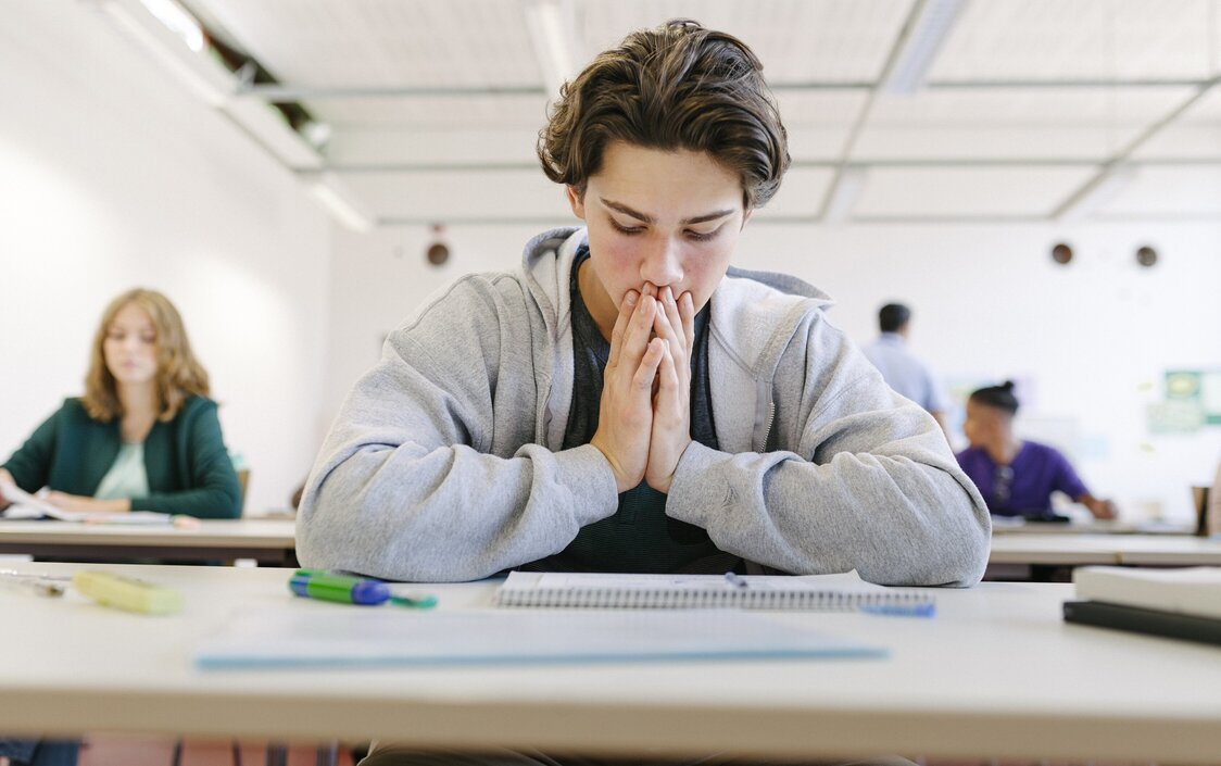 Ein Junge sitzt in der Schule am Tisch und guckt verzweifelt auf seine Unterlagen.