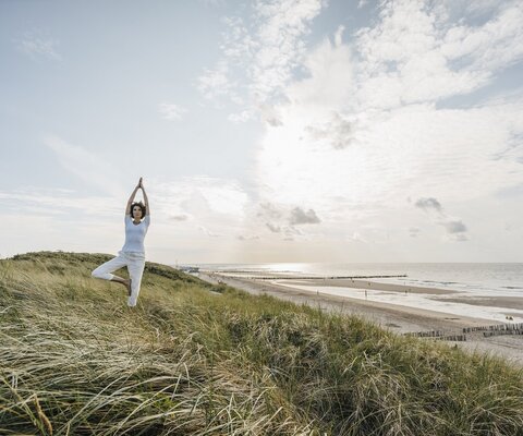 Eine Frau macht in den Dünen am Strand eine Yogaübung.