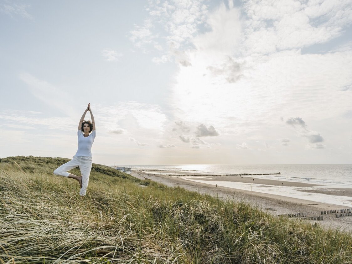 Eine Frau macht in den Dünen am Strand eine Yogaübung.