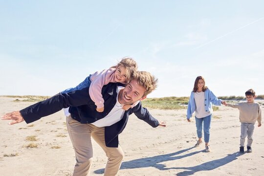 Ein Vater ht seine Tochter am Strand huckepack und seine Frau und sein Sohn laufen dahinter.