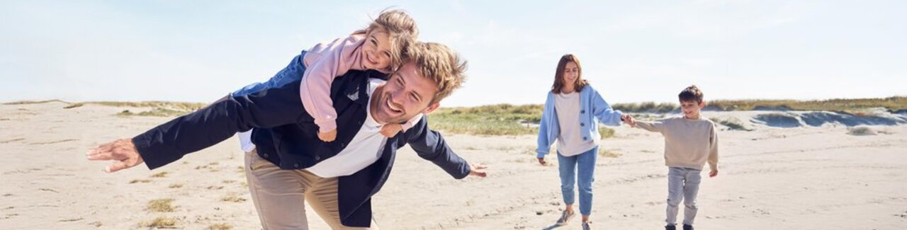 Ein Vater ht seine Tochter am Strand huckepack und seine Frau und sein Sohn laufen dahinter.