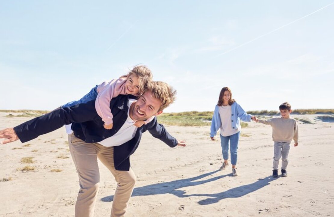 Ein Vater ht seine Tochter am Strand huckepack und seine Frau und sein Sohn laufen dahinter.
