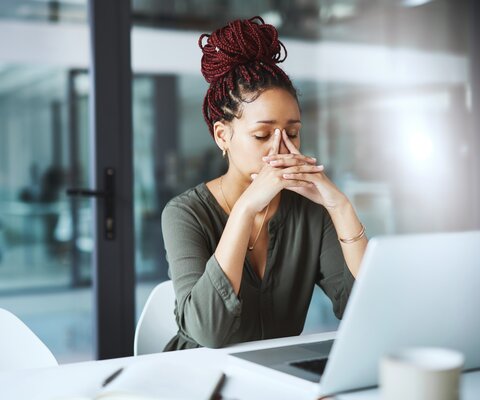 Eine Frau sitzt mit geschlossenen Augen vor ihrem Laptop am Schreibtisch. Sie reibt sich mit den Zeigefingern über den Nasenrücken.