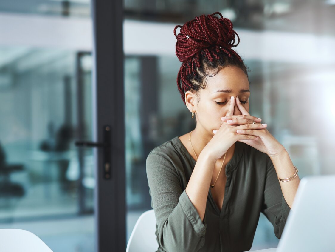 Eine Frau sitzt mit geschlossenen Augen vor ihrem Laptop am Schreibtisch. Sie reibt sich mit den Zeigefingern über den Nasenrücken.