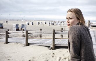 Eine Frau lehnt sich gegen das Geländer an einem Steg und guckt auf den Strand mit Strandkörben.