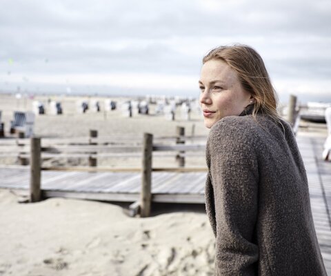 Eine Frau lehnt sich gegen das Geländer an einem Steg und guckt auf den Strand mit Strandkörben.