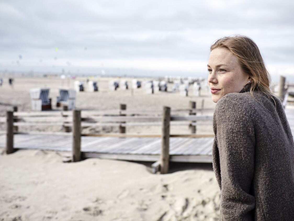 Eine Frau lehnt sich gegen das Geländer an einem Steg und guckt auf den Strand mit Strandkörben.
