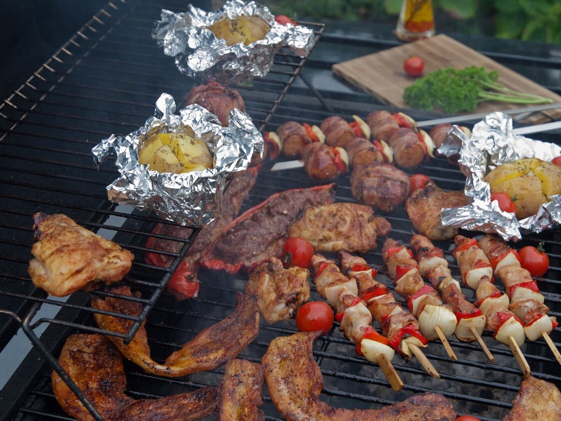 Auf einem Grill liegen Kartoffeln in Alufolie, Fleisch und Gemüsespieße.