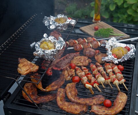 Auf einem Grill liegen Kartoffeln in Alufolie, Fleisch und Gemüsespieße.