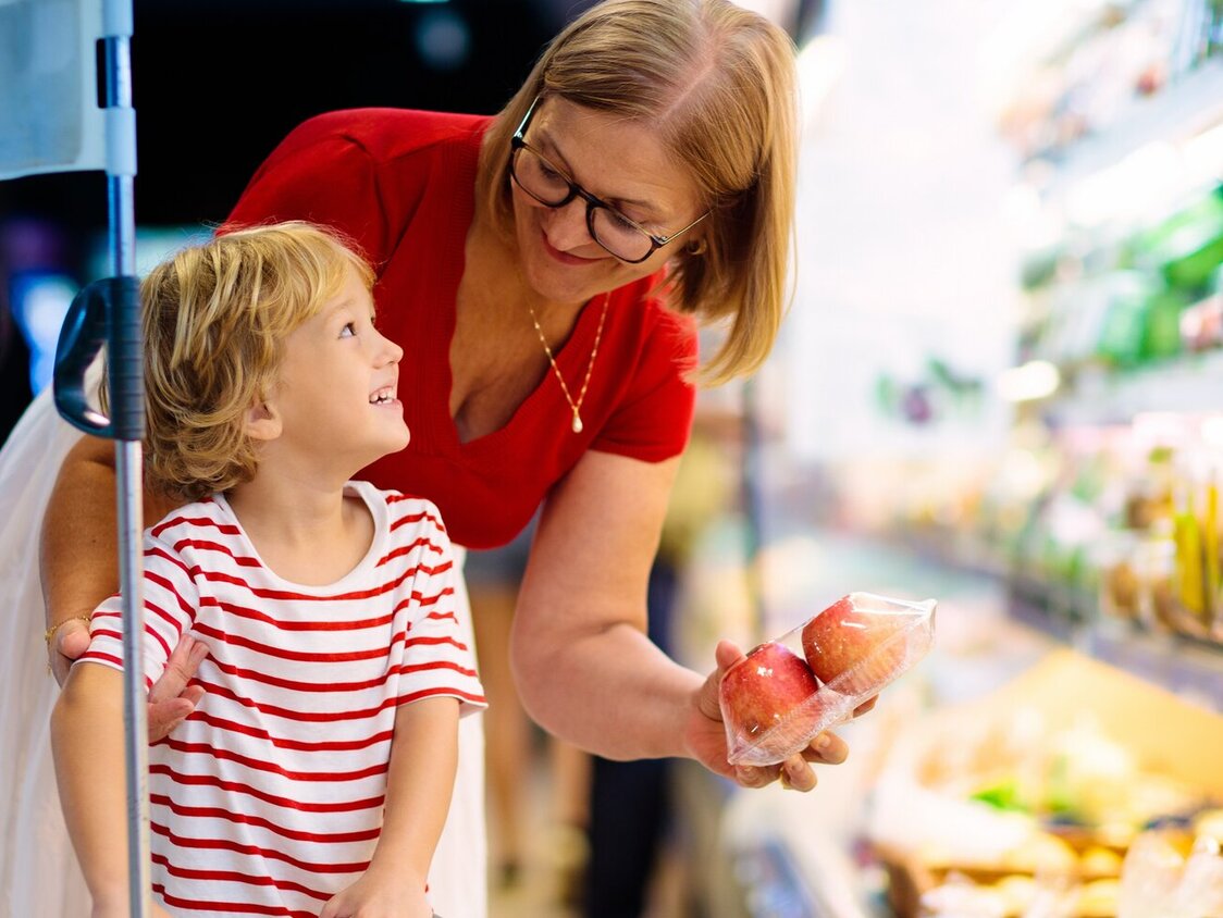 Eine Mutter, welche mit ihrem Kind vor einem Obstregal steht. Sie nimmt eine Packung Äpfel heraus und zeigt sie lächelnd dem Kind.