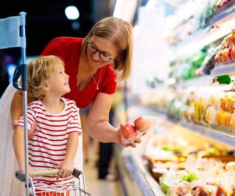 Eine Mutter, welche mit ihrem Kind vor einem Obstregal steht. Sie nimmt eine Packung Äpfel heraus und zeigt sie lächelnd dem Kind.