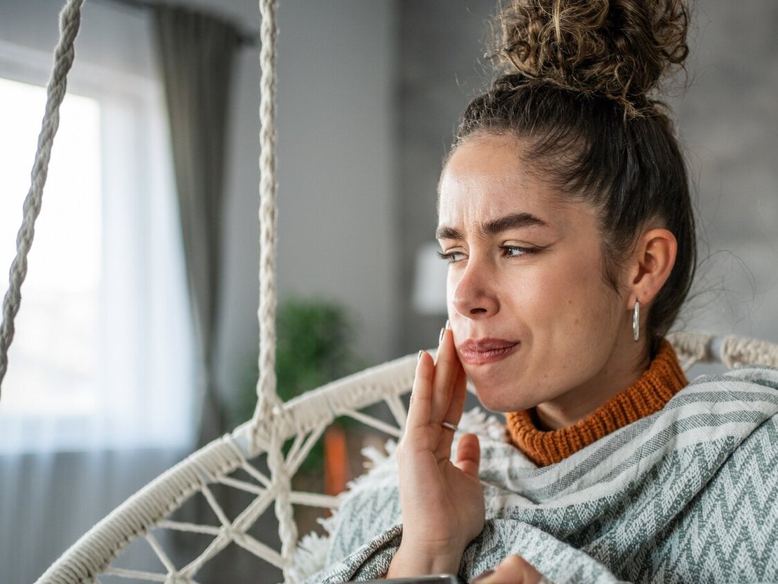 Junge Frau mit einer Tasse in der Hand, hält sich schmerzverzogen die Wange. Sie sitzt auf einer Schaukel im heimischen Wohnzimmer.