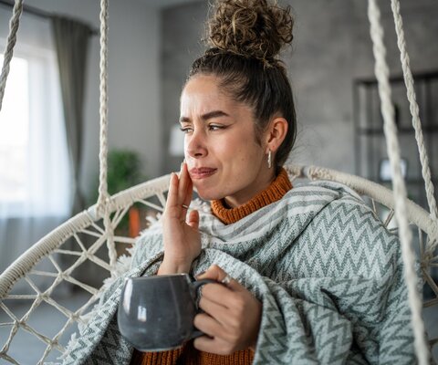 Junge Frau mit einer Tasse in der Hand, hält sich schmerzverzogen die Wange. Sie sitzt auf einer Schaukel im heimischen Wohnzimmer.
