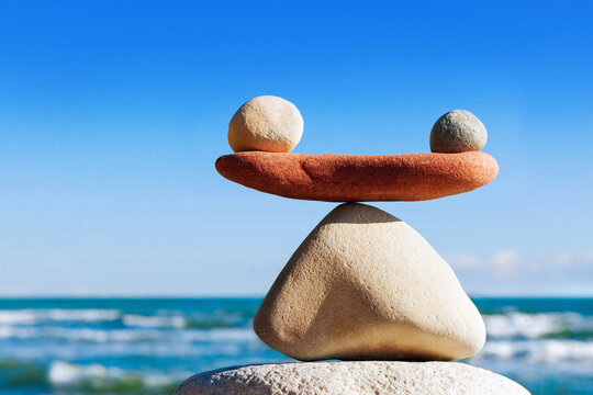 Zwei Steine sind zu einer Waage geformt. Rechts und links auf der Waage zwei weitere Steine. Im Hintergrund das Meer.