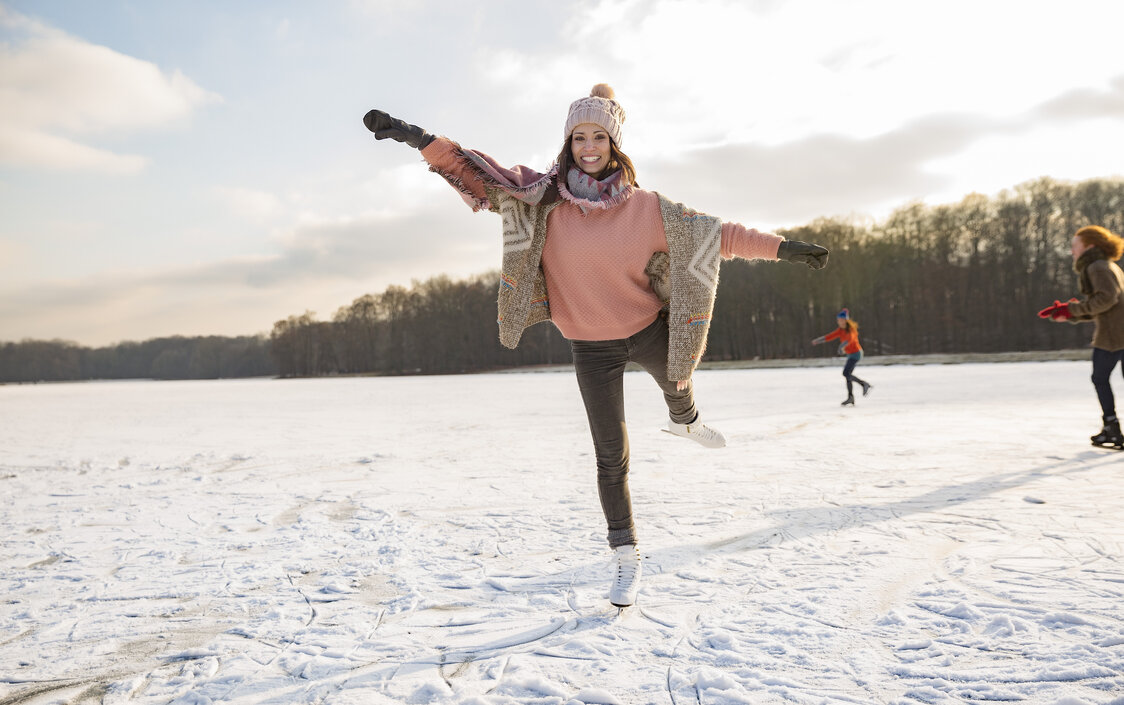 Eine Frau gleitet auf Schlittschuhen über eine Eislauffläche mit Menschen und Wald im Hintergrund.