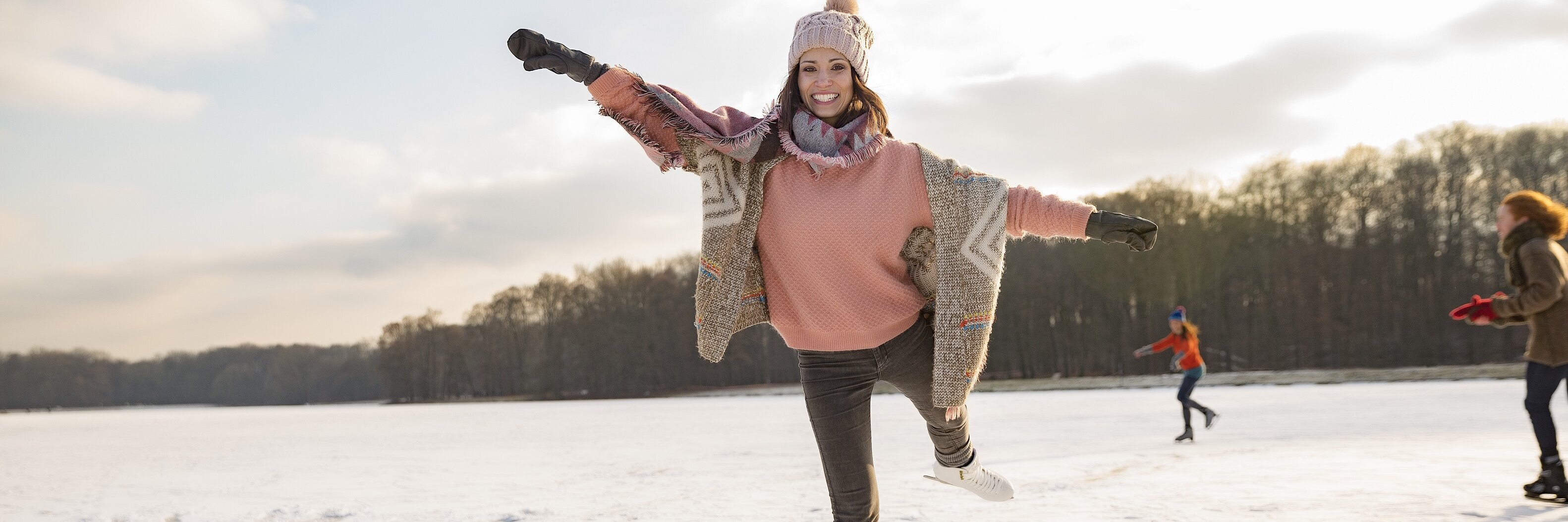 Eine Frau gleitet auf Schlittschuhen über eine Eislauffläche mit Menschen und Wald im Hintergrund.