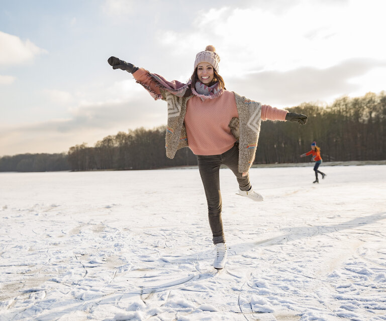 Eine Frau gleitet auf Schlittschuhen über eine Eislauffläche mit Menschen und Wald im Hintergrund.