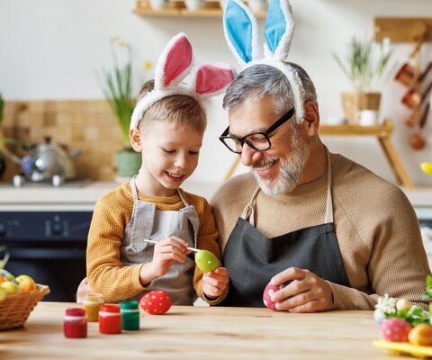 In einer Küche malt ein Kind mit seinem Opa Ostereier an. Beide tragen Hasenohren.