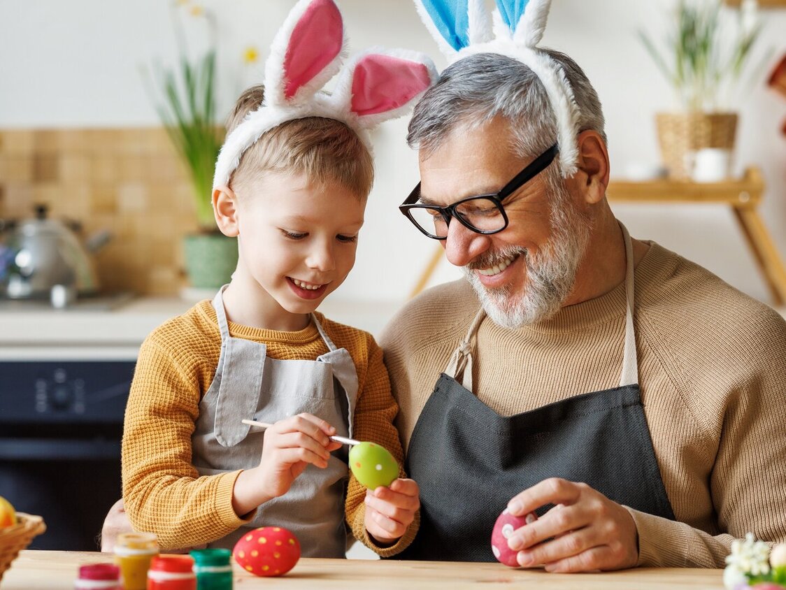 In einer Küche malt ein Kind mit seinem Opa Ostereier an. Beide tragen Hasenohren.