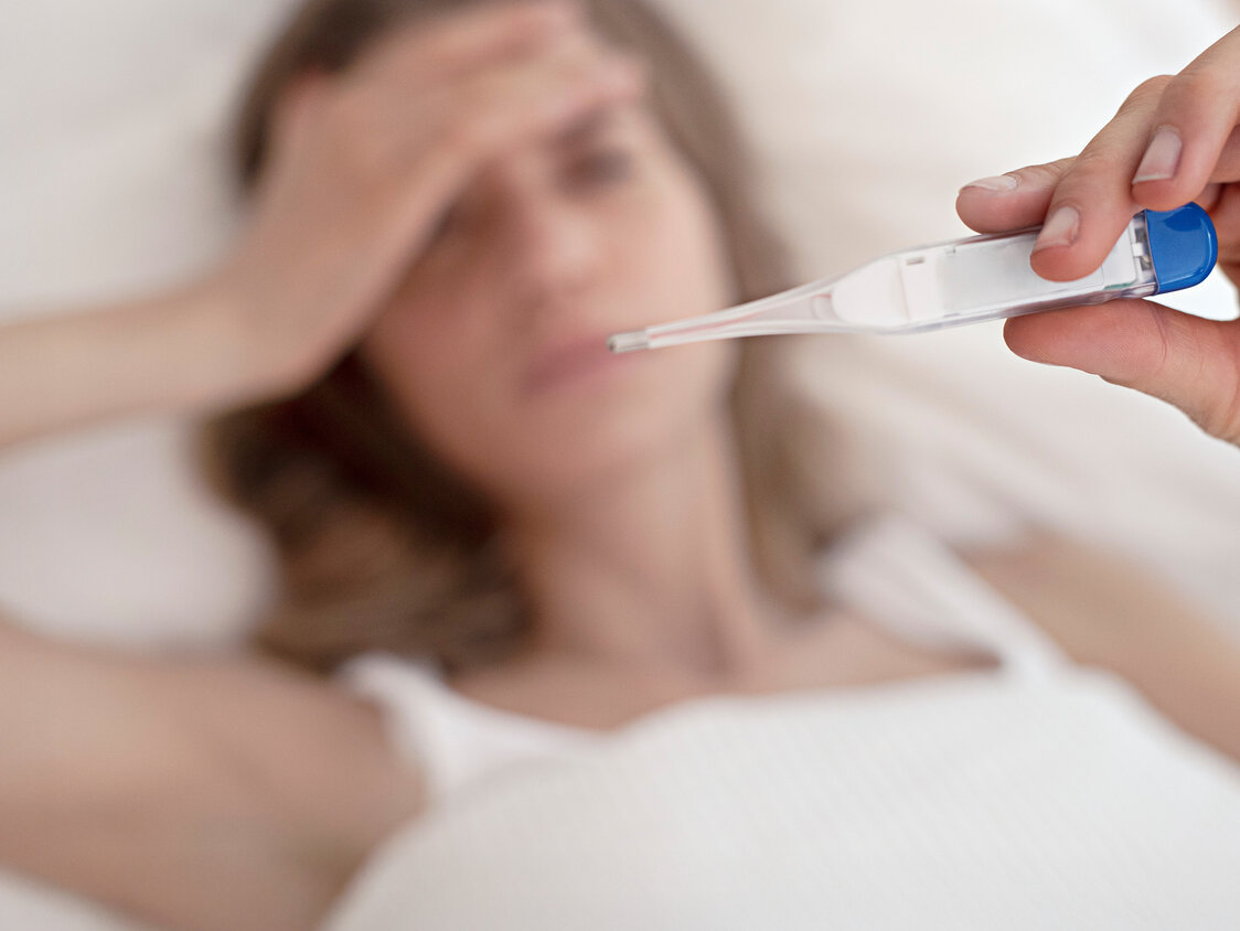 Eine liegende Frau hält ein Thermometer mit der Hand in die Luft, während die andere Hand auf ihrer Stirn liegt.